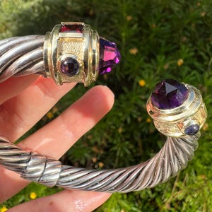 May include: A silver-tone cuff bracelet with gold accents and large, faceted gemstones. The bracelet features a twisted cable design and is adorned with a large, purple gemstone and smaller red and purple stones.