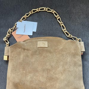 Może przedstawiać: Brązowa zamszowa torebka z łańcuchem w kolorze złotym. Torebka ma prostokątny kształt z zakrzywioną g&oacute;rą i małym złotym zapięciem. Nazwa marki "Nanushka" jest widoczna na torebce i na dołączonych metkach.