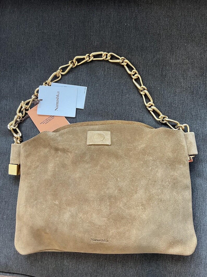 Może przedstawiać: Brązowa zamszowa torebka z łańcuchem w kolorze złotym. Torebka ma prostokątny kształt z zakrzywioną g&oacute;rą i małym złotym zapięciem. Nazwa marki "Nanushka" jest widoczna na torebce i na dołączonych metkach.