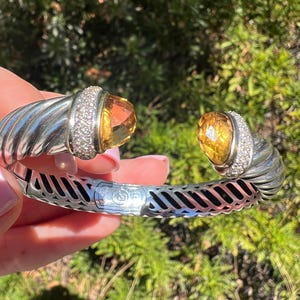 May include: A silver cuff bracelet with a twisted design. The bracelet features two large, faceted, yellow-orange gemstones, each surrounded by a ring of small, clear stones. The bracelet is held against a blurred green background.
