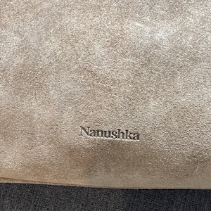 Może przedstawiać: Zbliżenie beżowego zamszu z wytłoczonym na powierzchni napisem "Nanushka". Tekstura wydaje się miękka i lekko zużyta, z subtelnymi odcieniami.