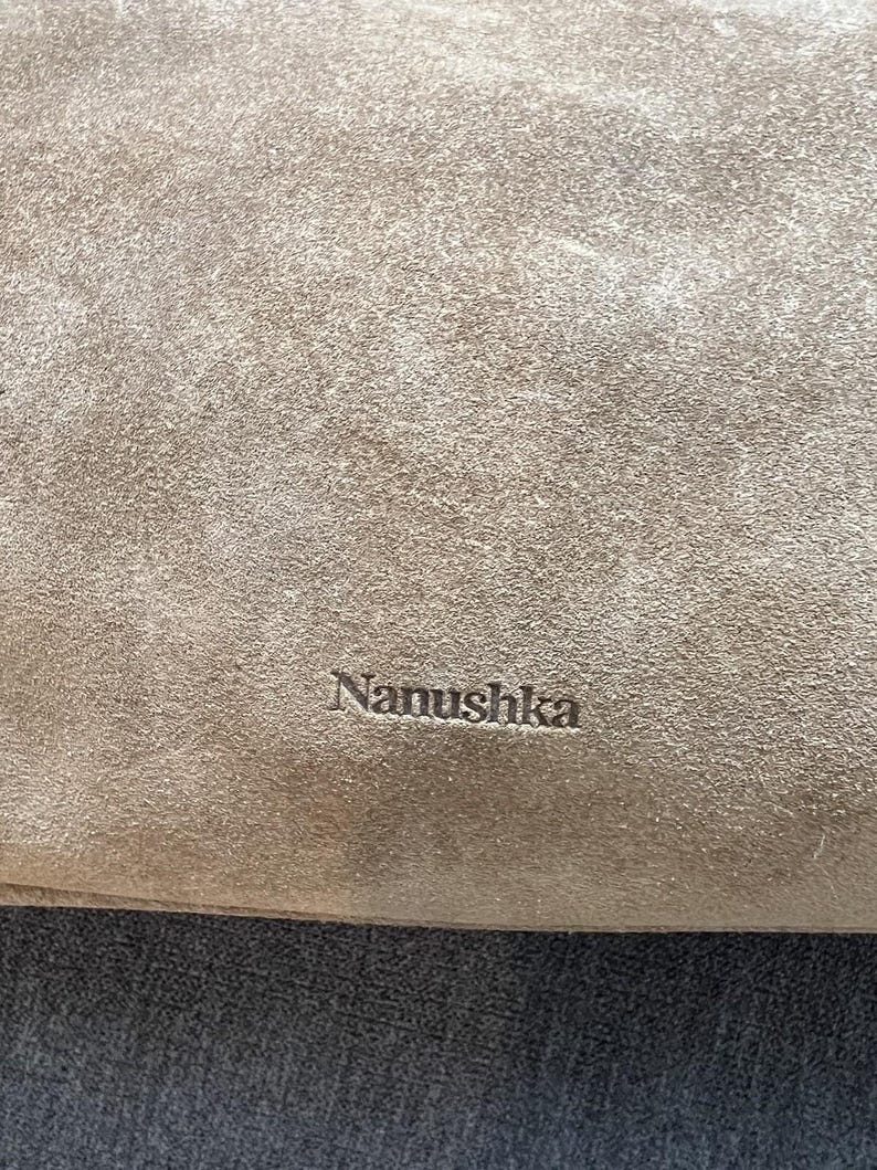 Może przedstawiać: Zbliżenie beżowego zamszu z wytłoczonym na powierzchni napisem "Nanushka". Tekstura wydaje się miękka i lekko zużyta, z subtelnymi odcieniami.