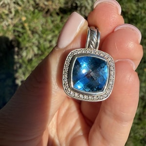 May include: A silver pendant with a large, square, faceted blue gemstone at its center. The gemstone is surrounded by a halo of small, clear gemstones. The pendant is held up by a silver bail with a twisted rope design.