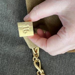 Może przedstawiać: Brązowa zamszowa torebka z łańcuszkiem w kolorze złotym. Torebka posiada kwadratowe, złote zapięcie z wytłoczonym wzorem. Łańcuch składa się z grubych, zazębiających się ogniw. Torebka jest trzymana przez dłoń.