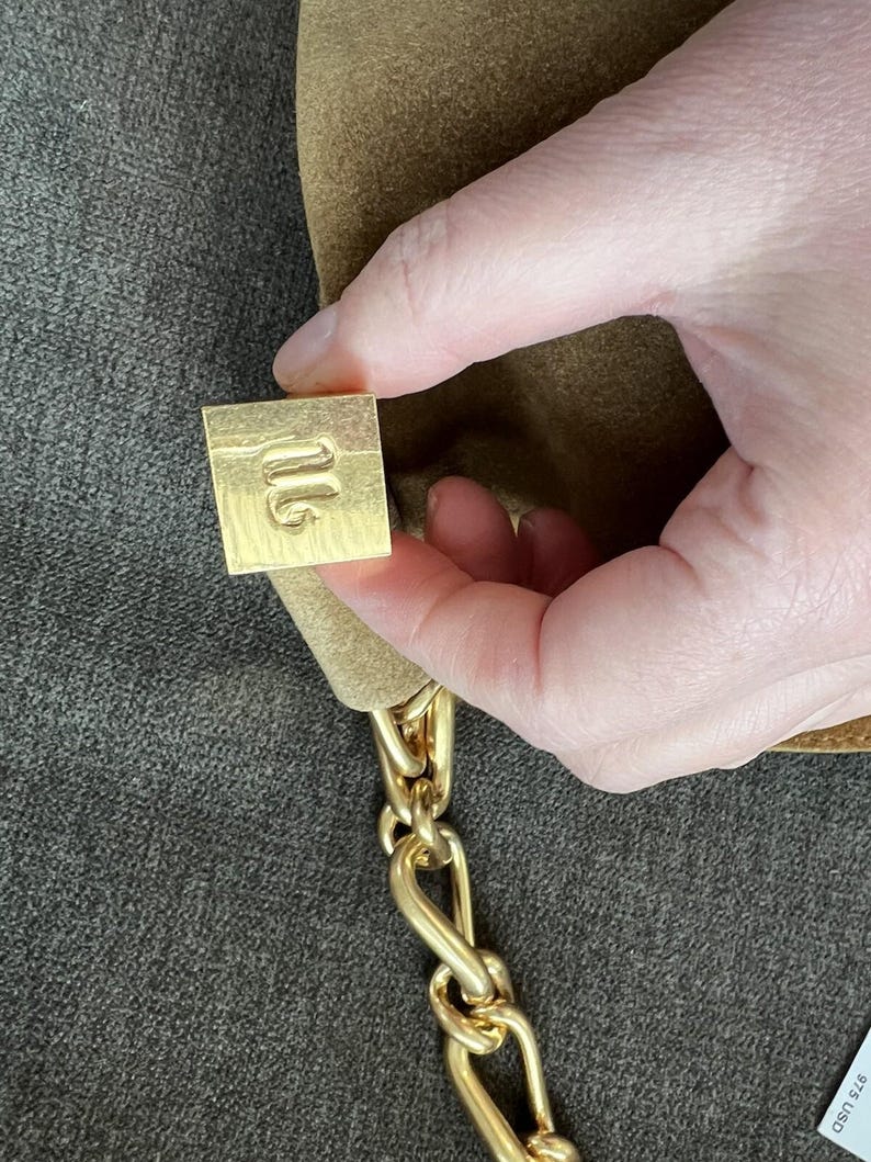 Może przedstawiać: Brązowa zamszowa torebka z łańcuszkiem w kolorze złotym. Torebka posiada kwadratowe, złote zapięcie z wytłoczonym wzorem. Łańcuch składa się z grubych, zazębiających się ogniw. Torebka jest trzymana przez dłoń.