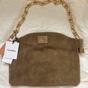 Może przedstawiać: Brązowa zamszowa torebka z łańcuchem w kolorze złotym. Torebka ma prostokątny kształt z zakrzywioną g&oacute;rą i małą, kwadratową naszywką z przodu. Etykieta marki głosi "Nanushka".
