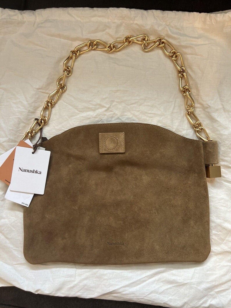 Może przedstawiać: Brązowa zamszowa torebka z łańcuchem w kolorze złotym. Torebka ma prostokątny kształt z zakrzywioną g&oacute;rą i małą, kwadratową naszywką z przodu. Etykieta marki głosi "Nanushka".