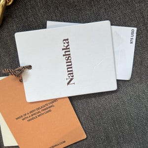 Może przedstawiać: Zbliżenie białych i beżowych etykiet produktu z nazwą marki "Nanushka" wydrukowaną na brązowo. Jedna etykieta zawiera tekst "MADE OF A VERY DELICATE FABRIC WITH A SPECIAL FINISHING, HANDLE WITH CARE." Na innej etykiecie widnieje napis "975 USD".