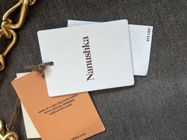 Może przedstawiać: Zbliżenie białych i beżowych etykiet produktu z nazwą marki "Nanushka" wydrukowaną na brązowo. Jedna etykieta zawiera tekst "MADE OF A VERY DELICATE FABRIC WITH A SPECIAL FINISHING, HANDLE WITH CARE." Na innej etykiecie widnieje napis "975 USD".