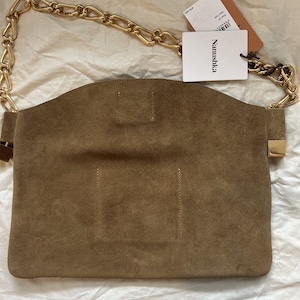 Może przedstawiać: Brązowa zamszowa torebka z łańcuchem w kolorze złotym. Torebka ma prostokątny kształt z zakrzywioną g&oacute;rą i złotym zapięciem. Dołączona jest biała metka z marką "Nanushka".