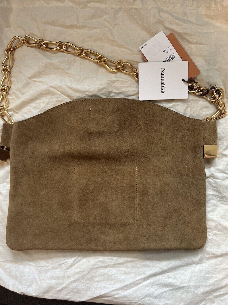 Może przedstawiać: Brązowa zamszowa torebka z łańcuchem w kolorze złotym. Torebka ma prostokątny kształt z zakrzywioną g&oacute;rą i złotym zapięciem. Dołączona jest biała metka z marką "Nanushka".