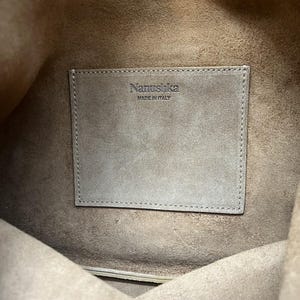 Może przedstawiać: Widok od wewnątrz brązowej sk&oacute;rzanej torby. Prostokątna naszywka z nazwą marki "Nanushka" i "MADE IN ITALY" jest wszyta wewnątrz. Wnętrze torby jest w jaśniejszym odcieniu brązu.