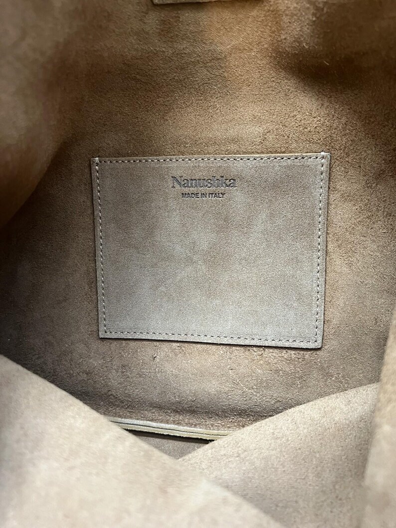 Może przedstawiać: Widok od wewnątrz brązowej sk&oacute;rzanej torby. Prostokątna naszywka z nazwą marki "Nanushka" i "MADE IN ITALY" jest wszyta wewnątrz. Wnętrze torby jest w jaśniejszym odcieniu brązu.