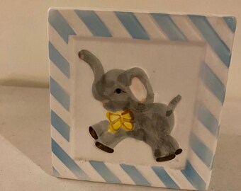 May include: A white ceramic piggy bank with a blue and white striped border. The bank features a hand-painted image of a gray elephant with a yellow bow tie.