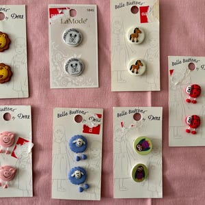 May include: A collection of decorative buttons featuring animal designs, including lions, horses, pigs, sheep, ladybugs, and owls. The buttons are attached to white cards with the brand names "Belle Buttons by Dritz" and "LaMode".