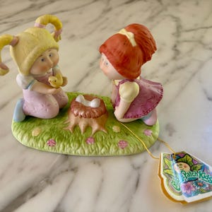May include: A ceramic figurine featuring two Cabbage Patch Kids. One child with blonde pigtails holds a yellow chick, while the other with red hair looks on. The base is a green lawn with a tree stump and flowers. A tag with the "Cabbage Patch Kids" logo is attached.
