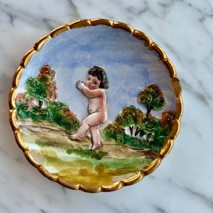 May include: A small, round ceramic plate with a gold rim and a raised, painted scene of a cherub in a landscape. The cherub is painted in shades of pink and white, and the landscape is painted in shades of green, blue, and brown.