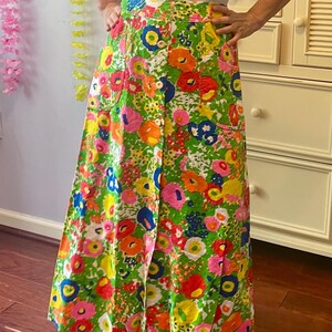 May include: A sleeveless white top with a floral print apron-style overlay and a long skirt. The skirt features a vibrant green background with large, colorful flowers in shades of pink, orange, yellow, and blue. The skirt has a button closure.