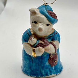May include: A ceramic figurine of a pig wearing a blue dress and hat, holding a baby pig. The figurine has a gold-colored bracelet and a maroon purse. A gold-colored wire loop is attached to the top of the hat.