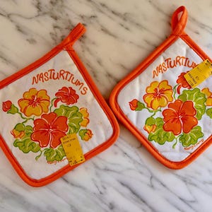 May include: Two square white fabric pot holders with orange trim and hanging loops. Each features a printed illustration of orange and yellow nasturtium flowers with green leaves and the text NASTURTIUMS. A yellow sale tag is attached to each.