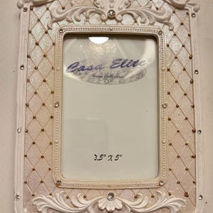 May include: White ornate picture frame with gold accents and rhinestones. The frame has a quilted pattern and the text "Casa Elite Home Collections" and "3.5" x 5" ".
