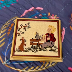 May include: Framed needlepoint art featuring a child with blonde hair, a dog, and a bird on a bench. The child wears a red jacket and gray pants. The frame is a distressed gold color with a red inner border. The artwork is on a cream-colored background.