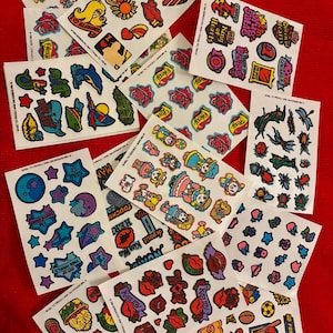 May include: A collection of vintage sticker sheets featuring various colorful designs. The stickers include images of hearts, stars, food items, and cartoon characters. Some stickers have text like "Sexy", "Love", and "Junk Food!"