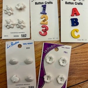 May include: Assortment of Le Bouton button crafts on white cards. Includes white teddy bear, bunny, and flower-shaped buttons, and colorful number and letter buttons. The cards are labeled with "WAL-MART" and prices.