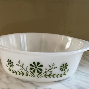 May include: White oval casserole dish with two handles. The dish features a green floral design around the exterior. The design includes stylized flowers and leaves. The dish is made of glass.