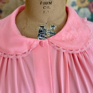 May include: A pink, long-sleeved nightgown with a lace trim collar and a button-up front. The collar has a floral embroidery design.