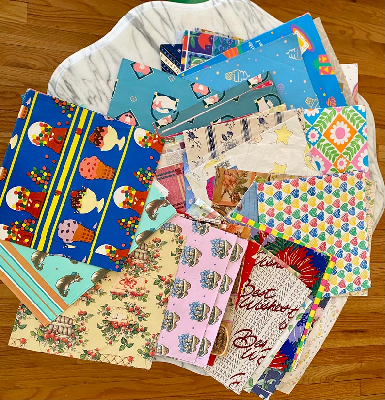 Peut inclure: Un assortiment de papiers d'emballage color&eacute;s avec diff&eacute;rents motifs, notamment des cornets de glace, des c&oelig;urs, des fleurs et des &eacute;toiles. Le papier est dispos&eacute; en pile sur une surface en marbre blanc.