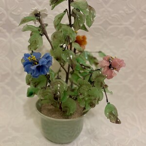 May include: A decorative glass flower arrangement in a green ceramic pot. The arrangement features colorful glass flowers in blue, pink, white, and orange, with green glass leaves. The pot is light green and has a textured design.