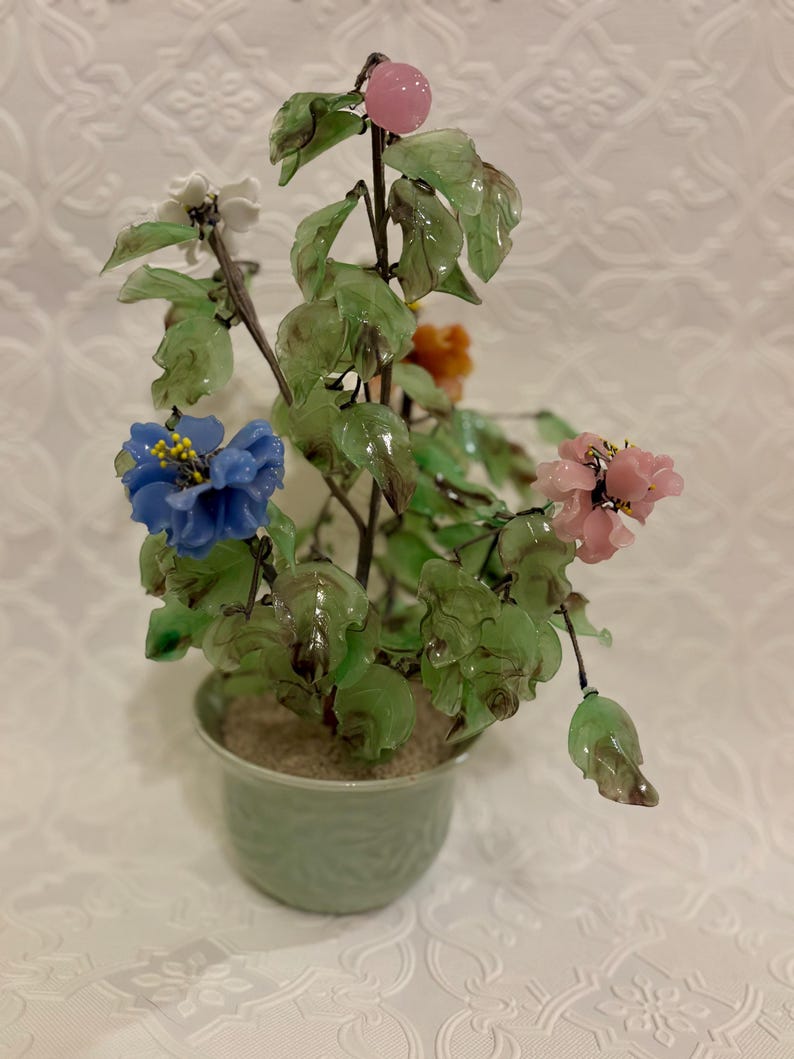 May include: A decorative glass flower arrangement in a green ceramic pot. The arrangement features colorful glass flowers in blue, pink, white, and orange, with green glass leaves. The pot is light green and has a textured design.