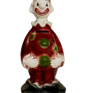 May include: A ceramic clown piggy bank with a red and white costume, green polka dots, and a black hat. The clown has a white face with a red smile and black eyes.
