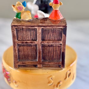 May include: A vintage ceramic music box featuring a brown cabinet with three cartoon figures on top. The cabinet sits on a yellow bowl with musical notes. The figures include a mouse, a black and white figure, and a figure in a red dress.