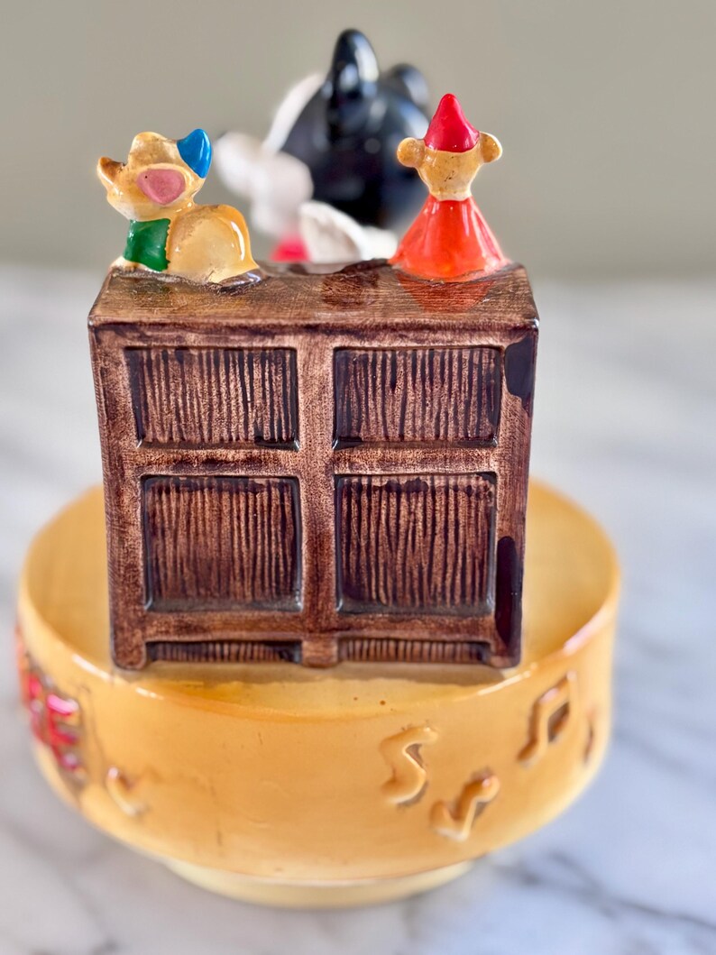 May include: A vintage ceramic music box featuring a brown cabinet with three cartoon figures on top. The cabinet sits on a yellow bowl with musical notes. The figures include a mouse, a black and white figure, and a figure in a red dress.