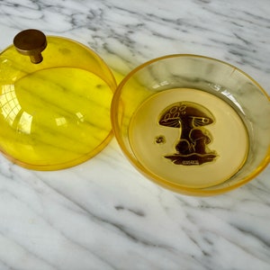 May include: A yellow plastic dome with a gold knob sits next to a yellow plastic dish with a gold mushroom design. The dish has the word "Shroomies" printed on it.