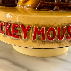 May include: A vintage ceramic figurine base with the words "MICKEY MOUSE" in red lettering. The base is a light tan color with a glossy finish. The figurine appears to be a music box base.