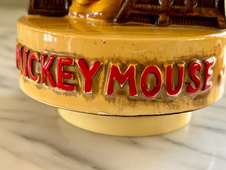 May include: A vintage ceramic figurine base with the words "MICKEY MOUSE" in red lettering. The base is a light tan color with a glossy finish. The figurine appears to be a music box base.