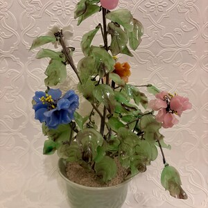 May include: A decorative glass flower arrangement in a green ceramic pot. The arrangement features blue, pink, white, and orange flowers, with green leaves and a pink fruit. The pot is light green and filled with a sandy material.