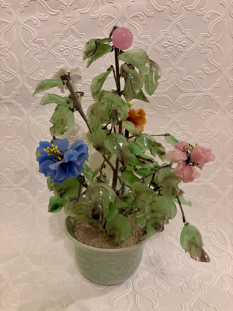 May include: A decorative glass flower arrangement in a green ceramic pot. The arrangement features blue, pink, white, and orange flowers, with green leaves and a pink fruit. The pot is light green and filled with a sandy material.