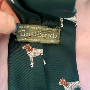 May include: A dark green necktie featuring a repeating pattern of brown and white spotted dogs. A rectangular fabric label is stitched to the back, reading: THE ORIGINAL DOG TIE COMPANY AT David-Barrato MANHASSET, N.Y. 11030.