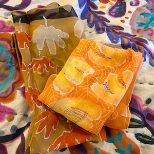 May include: Two sheer scarves with abstract floral patterns in shades of orange, yellow, and brown. The scarves are folded and layered on top of each other.