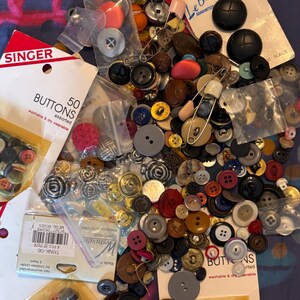 May include: A large collection of assorted buttons in various shapes, sizes, and colors, including black, red, silver, and brown. Some buttons are loose, while others are packaged in plastic bags or on branded Singer and Le Bouton cardboard cards.