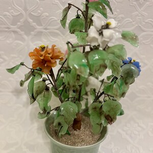 May include: A decorative potted plant with colorful glass flowers. The flowers are orange, white, and blue, with green leaves. The plant is in a light green pot filled with small pebbles.