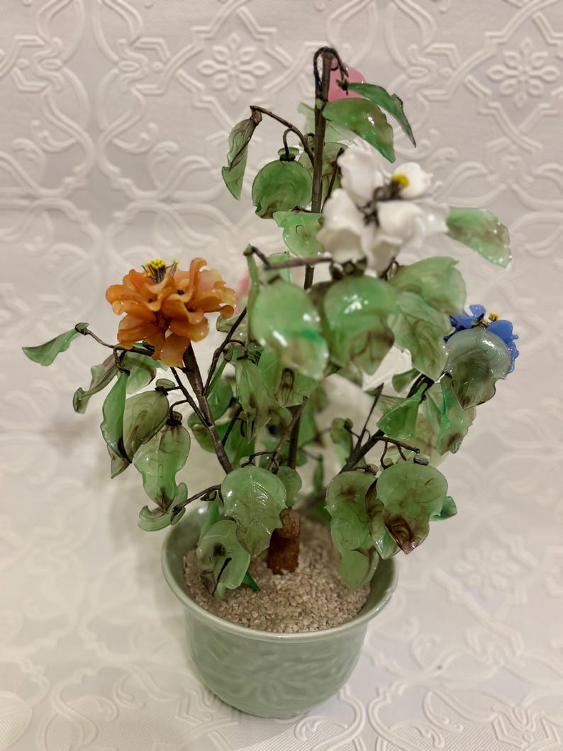 May include: A decorative potted plant with colorful glass flowers. The flowers are orange, white, and blue, with green leaves. The plant is in a light green pot filled with small pebbles.