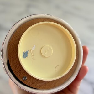 May include: A vintage music box base with a light yellow plastic top and a wooden base. The ceramic rim has a floral design. The text "JAPAN" is visible on the plastic top. A sticker with text is also visible.