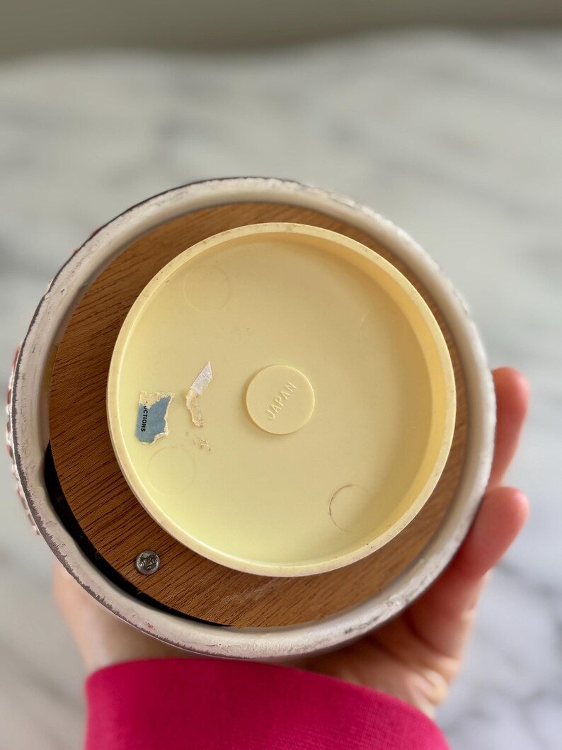 May include: A vintage music box base with a light yellow plastic top and a wooden base. The ceramic rim has a floral design. The text "JAPAN" is visible on the plastic top. A sticker with text is also visible.