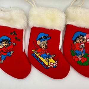 May include: Three red Christmas stockings with white faux fur trim. Each stocking features a cartoon mouse in a blue hat and red shirt. One mouse is sledding, one is holding a candy cane, and one is dancing.
