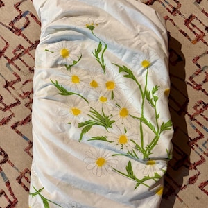 May include: White bed sheet set with a daisy floral pattern. The daisies have yellow centers and white petals, with green stems and leaves. The sheet set is folded and appears to be made of a soft fabric.